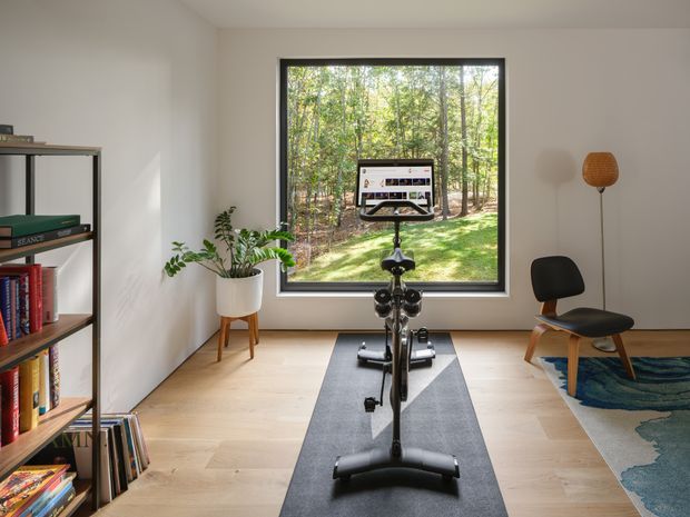 Bright, minimalist home gym viewed straight on, with a stationary exercise bike centered on a dark workout mat. The room has pale timber flooring, white walls, a large black-framed window overlooking a wooded landscape, open shelving with books and records, a potted plant, and a sculptural lounge chair with a floor lamp. The space feels calm, airy, and contemporary.