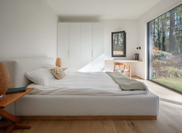 A bright, minimalist bedroom with a low upholstered platform bed, white bedding, built-in full-height white wardrobes, and pale timber flooring. A compact timber desk and chair sit beside a large floor-to-ceiling window overlooking a wooded landscape. Warm woven bedside lighting and a small neutral cushion add subtle texture.