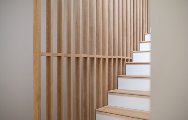 A minimalist interior stairway with light timber-tread stairs and white risers, bordered by a tall vertical timber slat screen and matching horizontal handrail or divider. The composition is clean and warm, with the stair rising toward a bright upper landing.