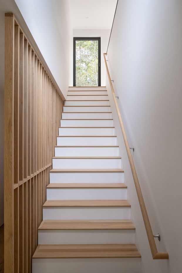 A bright, narrow residential stairway viewed looking upward. The staircase has white risers and light timber treads, a simple timber handrail with metal brackets, and a full-height vertical timber slat balustrade along the left side. A tall dark-framed window at the landing brings in natural light and views of trees. The space is clean and minimalist with white walls and ceiling.