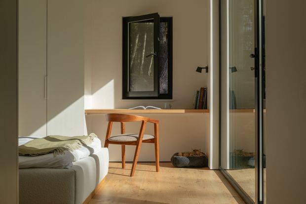 A compact, softly lit bedroom corner with a low white upholstered bed or sofa at left, a slim timber writing desk and mid-century-style wooden chair beneath a small black-framed window, and full-height mirrored wardrobe doors on the right. Pale walls, light timber flooring, and restrained furnishings create a warm, calm, minimalist interior.