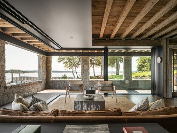 A spacious indoor-outdoor living room with exposed timber ceiling beams, dark-framed expansive glazing, and rustic stone walls and columns. A large sofa faces a stone coffee table and two lounge chairs, with views toward a lawn and waterfront beyond. The room has a warm, relaxed, natural character and strong visual connection to the outdoors.