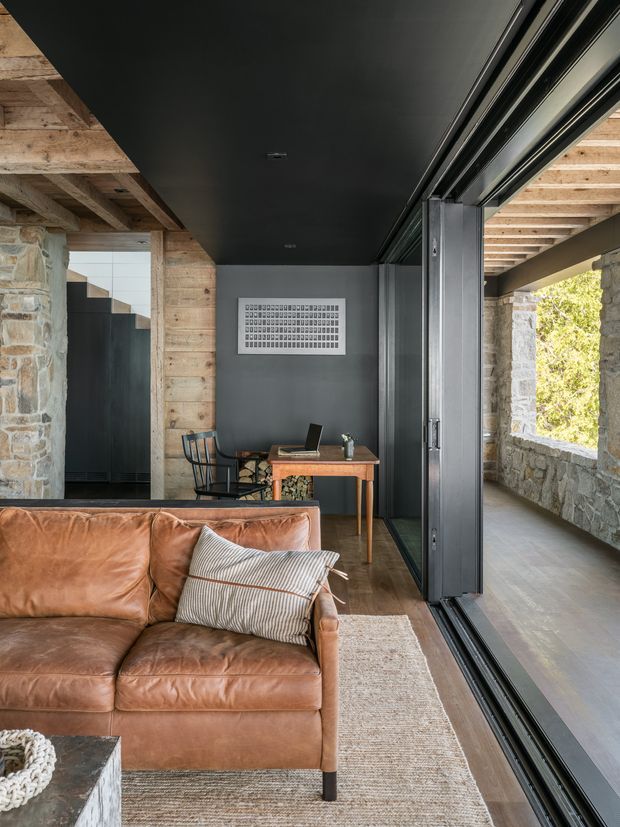 A compact, refined living room and home office corner with a cognac leather sofa in the foreground, a timber desk and chair against a dark wall, and extensive black-framed sliding glazing opening to a stone-lined exterior walkway. The interior combines exposed timber ceiling beams, rough natural stone walls, dark ceiling and wall finishes, timber flooring, and a textured neutral rug. The space has a rustic-modern, warm yet moody character with strong indoor-outdoor connection.