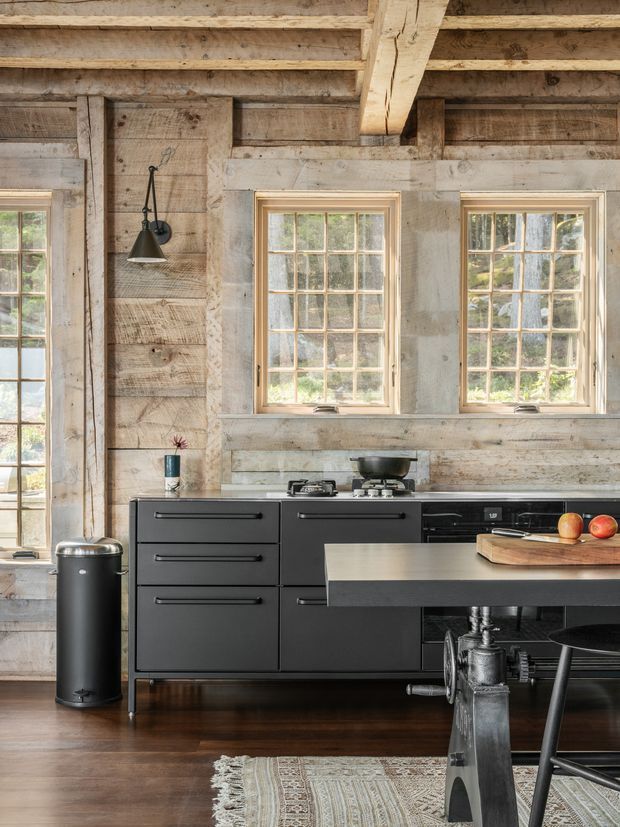 Rustic kitchen interior with exposed reclaimed timber wall and ceiling structure, multiple divided-light windows, matte black cabinetry and appliances, and a dark island bench with industrial metal supports. Dark timber flooring, a woven rug, and simple black pendant lighting create a warm, characterful farmhouse-industrial atmosphere.