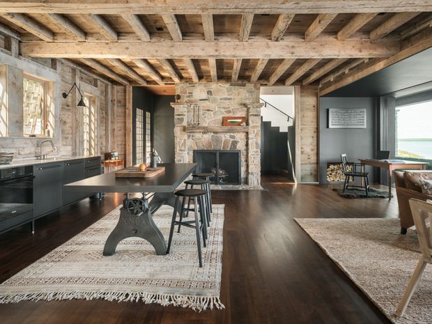 Open-plan rustic interior combining a kitchen, dining area and living space. The room features exposed timber ceiling beams and boards, weathered timber wall cladding, a substantial natural-stone fireplace, dark kitchen cabinetry, a dark wood floor, and large windows overlooking a bright exterior view. A long island or dining table with industrial metal supports sits centrally, while a dining table and chairs are positioned near the fireplace and glazing.
