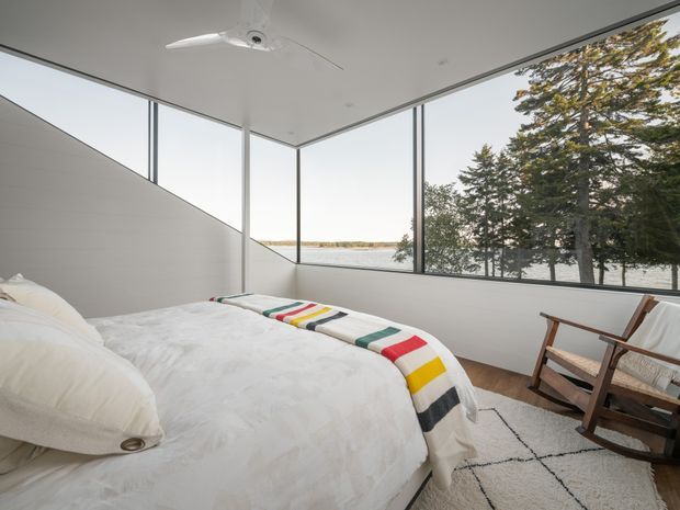 A bright, minimalist bedroom with a low-profile bed dressed in white bedding and a colourful striped throw. Full-height corner glazing overlooks a calm body of water and trees, while a timber rocking chair sits on a pale textured rug. A white ceiling fan and clean-lined wall finishes reinforce the simple contemporary aesthetic.