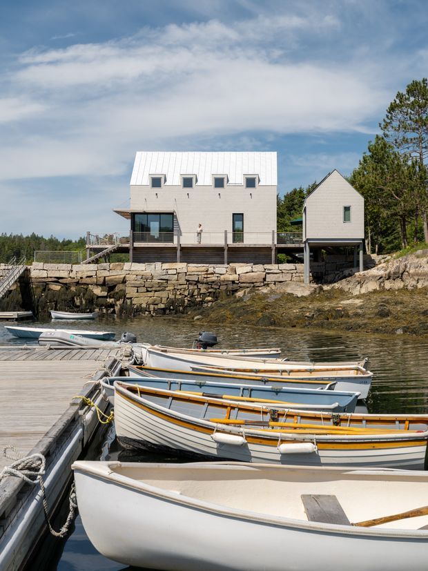 Exterior view of a contemporary coastal house composed of two raised, light-coloured volumes on a rocky waterfront site. The main rectangular building has a standing-seam metal roof, horizontal light cladding, small upper windows, large glazed doors, and a deck. A smaller gabled volume stands to the right on supports. Stone retaining walls, timber decking, dock structures, and several small boats occupy the foreground.