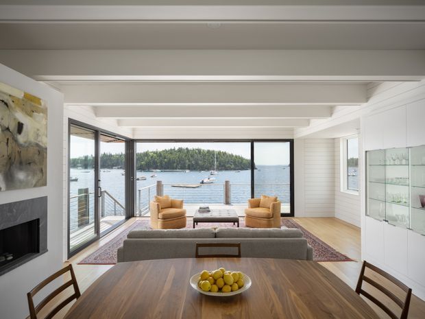 Bright open-plan living and dining space with a waterfront view through expansive black-framed glazing. The room features white painted walls and ceiling beams, horizontal wall panelling, warm timber flooring and dining furniture, a fireplace, a grey sofa, mustard armchairs, and built-in glass display cabinetry. A deck and calm water are visible beyond the sliding doors.