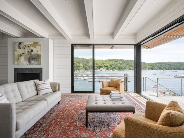 Bright waterfront living room with expansive black-framed glazing opening onto a deck and views across a sheltered bay. The room features white horizontal wall panelling, a white coffered and beamed ceiling, a dark fireplace surround, a large pale upholstered sofa, woven lounge chairs, a tufted ottoman, and a richly patterned traditional rug.