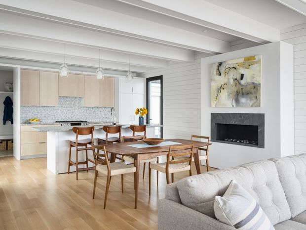 Bright open-plan coastal-inspired interior combining a kitchen, dining area and living room. The space features white-painted timber wall panelling, exposed white ceiling beams, pale timber flooring, light wood kitchen cabinetry, a patterned tile splashback, a dark fireplace surround, and mid-century-style timber dining furniture. Large abstract artwork and a light upholstered sofa add softness to the refined, airy room.