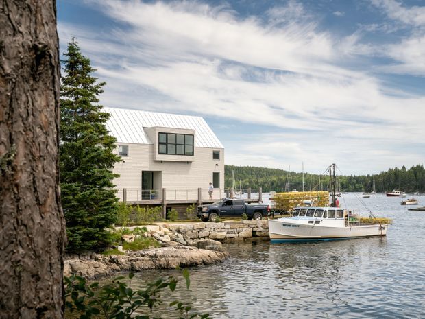 A modern light-coloured waterfront building with a white standing-seam pitched roof and projecting upper-level glazing, positioned directly beside a rocky shoreline and small marina. A dock, fishing boat, vehicle, evergreen trees, and forested shoreline are visible under a bright partly cloudy sky.