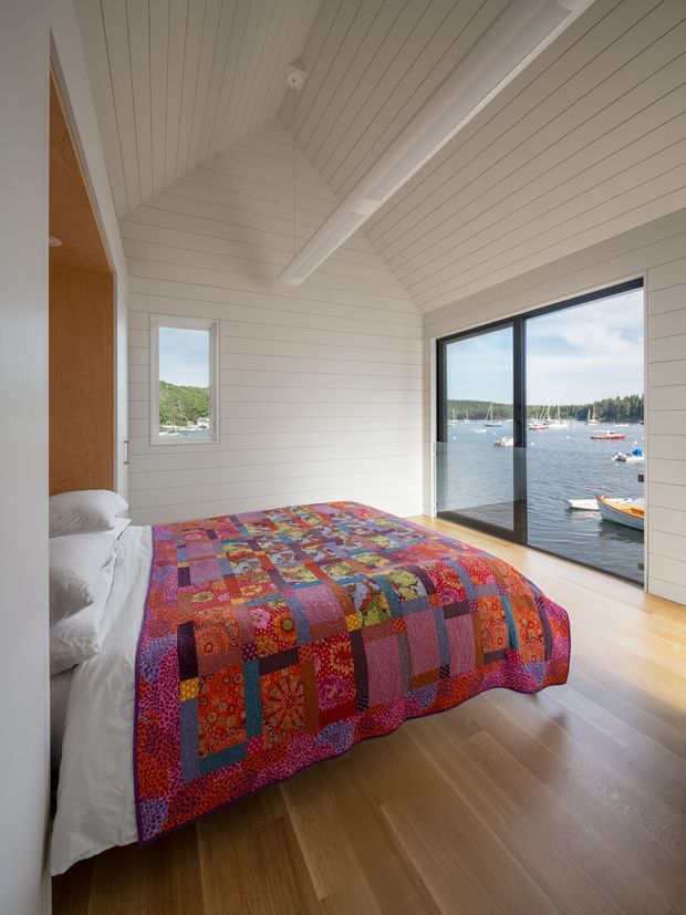 A bright bedroom with a low pitched, vaulted ceiling clad in white painted horizontal boards, exposed white structural beams, white wall panelling, and warm timber flooring. A bed with a colourful patchwork quilt occupies the room, while large dark-framed glazing provides a direct view over a calm harbour with boats and wooded shoreline. A timber-toned built-in surface is visible beside the bed.