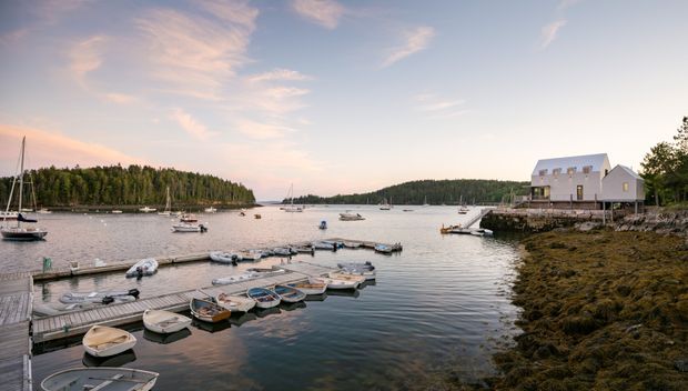 A tranquil coastal waterfront scene viewed from a dock, with floating timber pontoons and small boats in calm water. A light-coloured gabled building stands on a rocky shoreline to the right, while wooded shorelines and moored boats extend into the distance under a softly lit sky.
