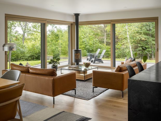 A bright, refined living room with expansive floor-to-ceiling timber-framed glazing overlooking a leafy garden. The space features a freestanding wood-burning stove, two cognac leather sofas, a low glass-and-timber coffee table, a textured grey rug, pale timber flooring, and a dark stone-clad architectural element in the foreground. A dining area is partially visible at left, while outdoor lounge chairs can be seen beyond the glazing.