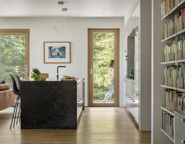 A bright, contemporary open-plan kitchen with a large dark stone island and slim black bar stools. Pale timber flooring, white cabinetry, integrated appliances, and full-height timber-framed glazing create a clean, warm interior. A built-in floor-to-ceiling bookcase lines the right wall, while a living area and garden views are visible beyond the kitchen.