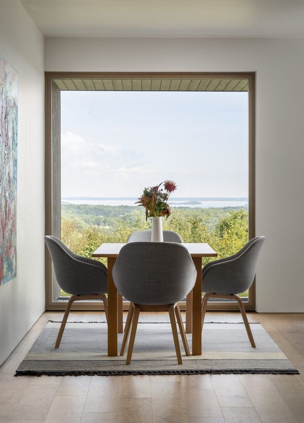 A serene dining area arranged in front of a large picture window overlooking a wooded landscape and distant water. The space features a simple rectangular timber dining table, four charcoal upholstered shell chairs with timber legs, a pale textured rug, light timber flooring, white walls, and a timber-framed window. A small vase of flowers sits centrally on the table, while a painted artwork is partially visible on the side wall.