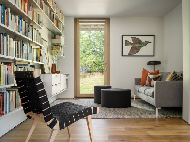 A bright, refined reading and living space with floor-to-ceiling built-in bookshelves along the left wall, a mid-century lounge chair in the foreground, and a compact grey sofa with patterned cushions. A low dark ottoman sits on a textured rug, while a glazed timber-framed door opens to greenery outside. The room has light timber flooring, white walls and joinery, a simple black floor lamp, and framed bird artwork.