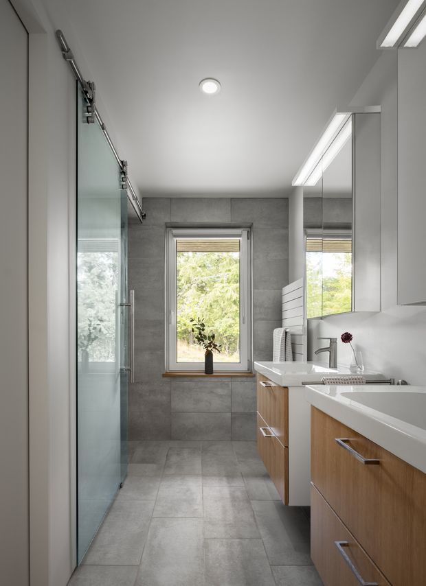 A bright, narrow contemporary bathroom with a walk-in glass shower along the left wall, a double vanity with white countertops and timber-look drawers on the right, and large grey stone-look tiles covering the floor and walls. A central window brings in natural greenery views, while mirrored cabinetry and linear ceiling lighting enhance the space.