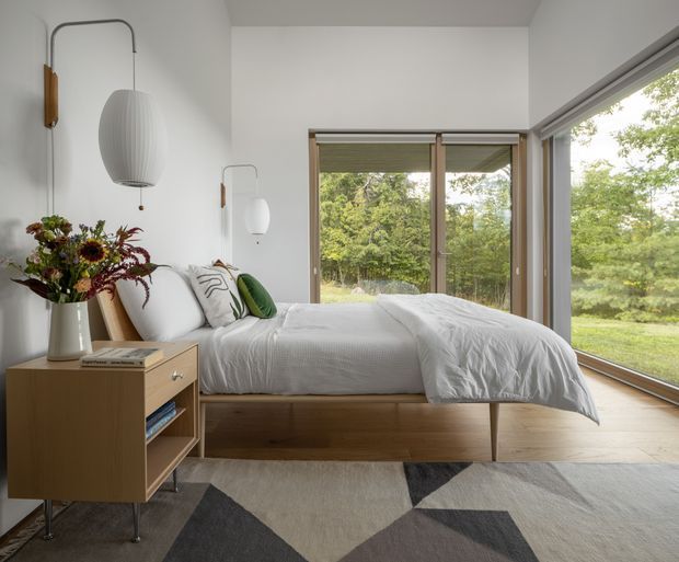 A bright, contemporary bedroom with a low platform bed in light timber, white bedding, and large corner windows overlooking greenery. A compact timber bedside cabinet, sculptural pendant and wall lights, patterned cushions, and a geometric grey, white, and charcoal rug add restrained mid-century-inspired detail.