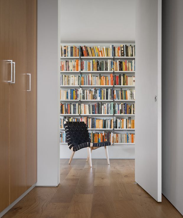 A minimalist reading nook viewed through a doorway, with a full-height wall of densely packed bookshelves and a black woven lounge chair positioned in front. The space has pale grey walls, warm timber flooring, and a tall timber built-in cabinet with simple white handles on the left. The clean, restrained composition creates a quiet library-like atmosphere.