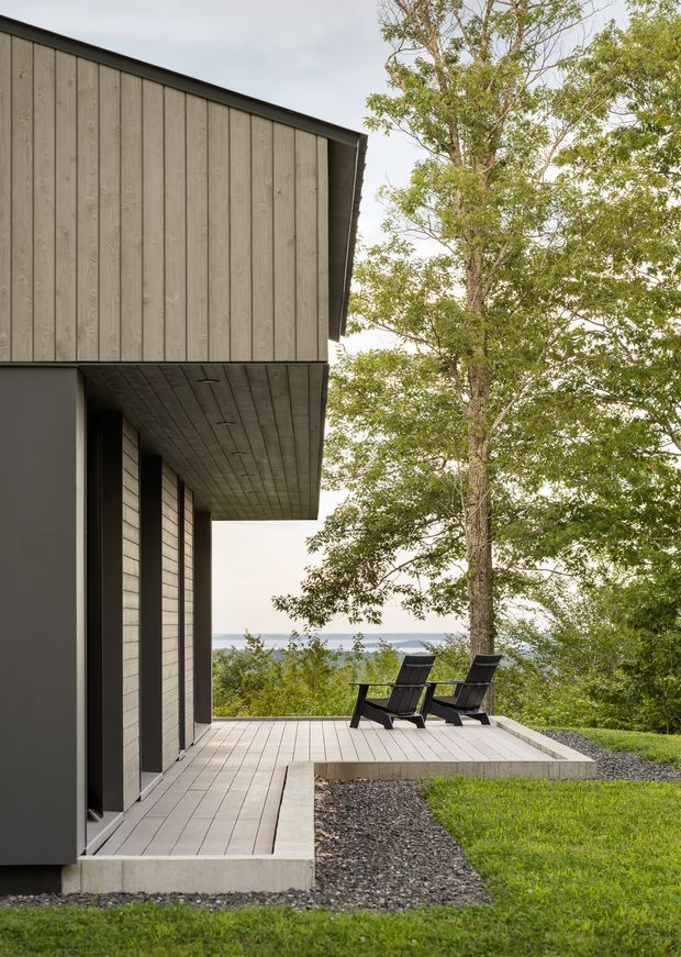A modern dark-clad house viewed from an elevated outdoor deck. The building has a projecting flat or very low-pitched roof with deep eaves, vertical weatherboard-style cladding, dark wall panels, and a timber-lined soffit. Two lounge chairs sit on a pale timber deck overlooking a wooded landscape and distant water or hills. The scene has a restrained, contemporary architectural character with strong indoor-outdoor connection.