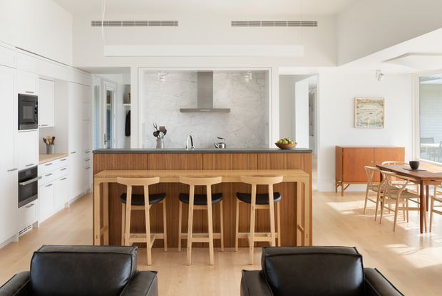 Bright open-plan kitchen and dining area viewed from the living room. The kitchen features white full-height cabinetry, integrated appliances, a large timber island with a dark benchtop, three timber bar stools, and a pale stone splashback with a simple metal rangehood. A light timber dining table and chairs occupy the adjoining space, while black leather seating is visible in the foreground. High ceilings, linear ceiling vents, and broad openings create an airy contemporary interior.