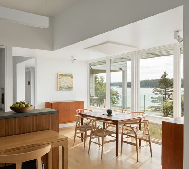 Bright open-plan dining area beside a kitchen, with a mid-century-style timber dining table and chairs, warm wood cabinetry, pale timber flooring, and expansive white-framed glazing overlooking a wooded waterfront. The room has a high sloping ceiling, crisp white walls, and abundant natural light.