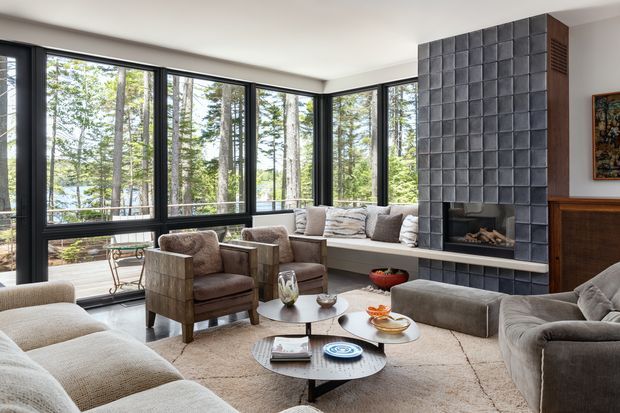 A refined living room with expansive floor-to-ceiling corner glazing overlooking a wooded setting. The room features a large sectional sofa, upholstered lounge chairs, layered round coffee tables, a textured area rug, and a prominent dark tiled fireplace wall with integrated firebox and hearth. Dark window frames and warm wood cabinetry add contrast to the neutral furnishings.