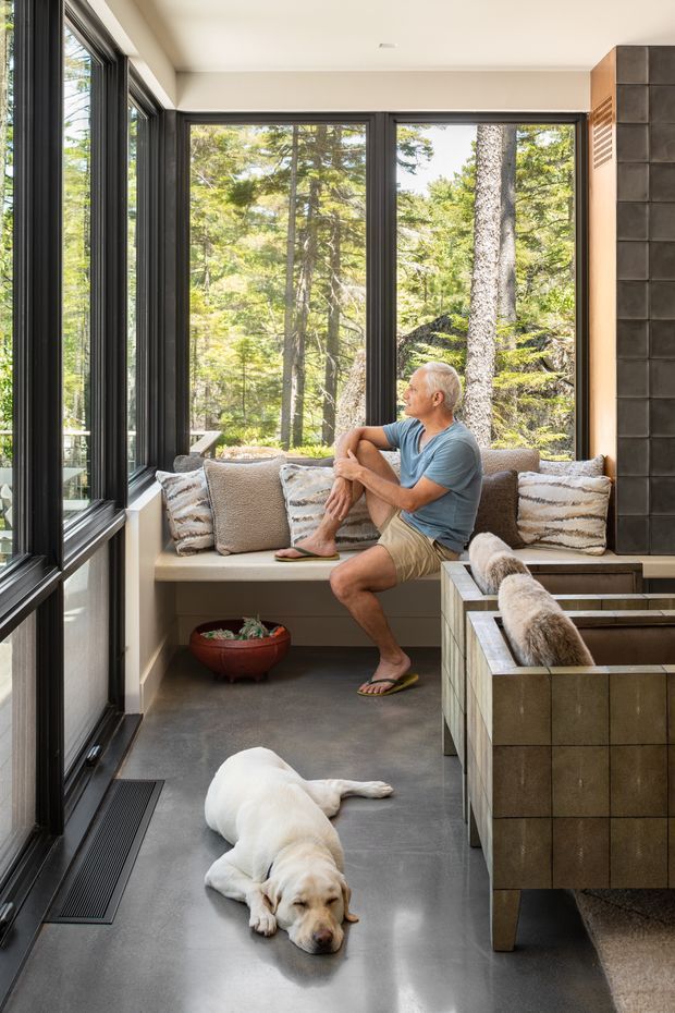 A bright, narrow sunroom or enclosed sitting area with floor-to-ceiling black-framed glazing on two sides, looking into a wooded landscape. A built-in upholstered window seat with cushions occupies the rear, while a tiled or stone-clad built-in bench or partition is visible on the right. The room has a smooth grey concrete-like floor, warm off-white walls and ceiling, and a relaxed seating area with a person and dog. The space feels airy, calm and connected to nature.
