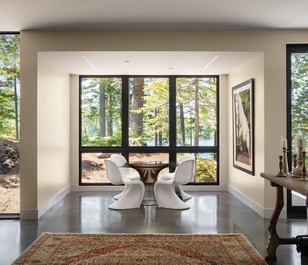 A bright, modern dining nook framed by large black window assemblies overlooking a wooded landscape. Four sculptural white dining chairs surround a compact round table, with polished grey flooring, warm off-white walls, recessed linear ceiling lights, and a traditional patterned area rug in the foreground. A framed artwork and a dark wood console with decorative objects sit along the right wall.