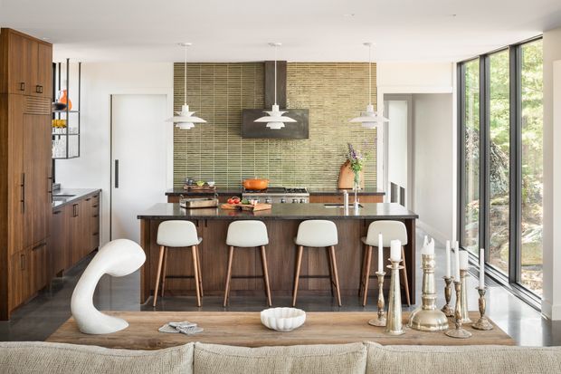 Bright contemporary kitchen viewed from an adjoining living area, featuring a large dark timber island with seating for four, full-height timber cabinetry, integrated appliances, pendant lights, a textured green-grey tiled backsplash, and expansive black-framed glazing along one side. A sculptural white chair and decorative objects sit in the foreground.