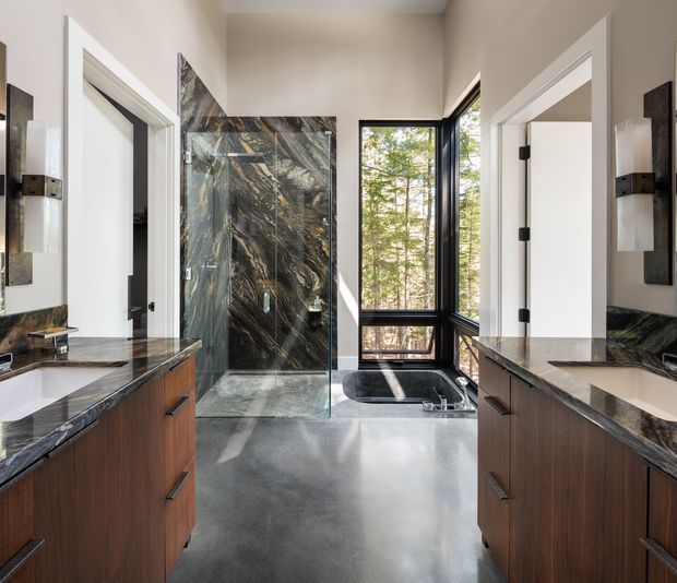 A contemporary bathroom viewed between two dark timber double vanities, with white undermount basins, dramatic veined dark stone benchtops and a matching full-height shower feature wall. A freestanding bathtub sits beside large black-framed windows overlooking trees, while pale walls, grey flooring and wall sconces create a refined, high-contrast interior.