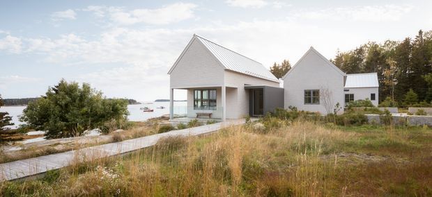 A contemporary coastal residential compound composed of several small white-clad gabled volumes. The main building has a steep pitched metal roof, vertical board-like light cladding, large glazed openings and a covered entry connection to a smaller adjoining structure. Pale timber or concrete paths cross a natural grassy site, with water visible beyond the house and trees in the background.