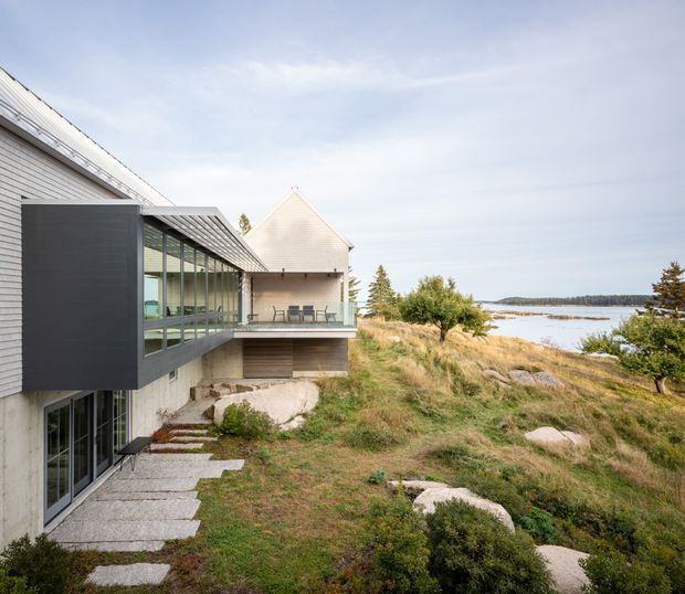 Exterior view of a contemporary coastal residence set among exposed granite rocks and natural grasses beside a calm body of water. The house combines light horizontal weatherboard cladding, a dark projecting enclosed volume, extensive glazed walls, and a projecting terrace beneath a steep gabled roof. The image is bright and airy, with the architecture integrated into the rocky shoreline landscape.