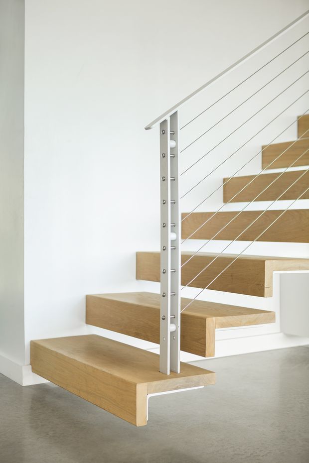 A minimalist open-riser staircase with thick light timber treads cantilevered from a white wall, paired with a slim white-painted metal post and horizontal cable balustrade. The staircase is set against white walls with a smooth grey concrete floor, creating a bright, clean contemporary interior.