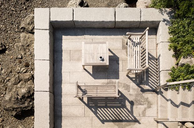 Overhead view of a compact stone-paved outdoor terrace enclosed by low pale masonry walls, with a timber slatted lounge chair, bench, and small square table. Strong sunlight creates crisp linear shadows from the furniture and nearby railing; rocks and planting border the terrace.