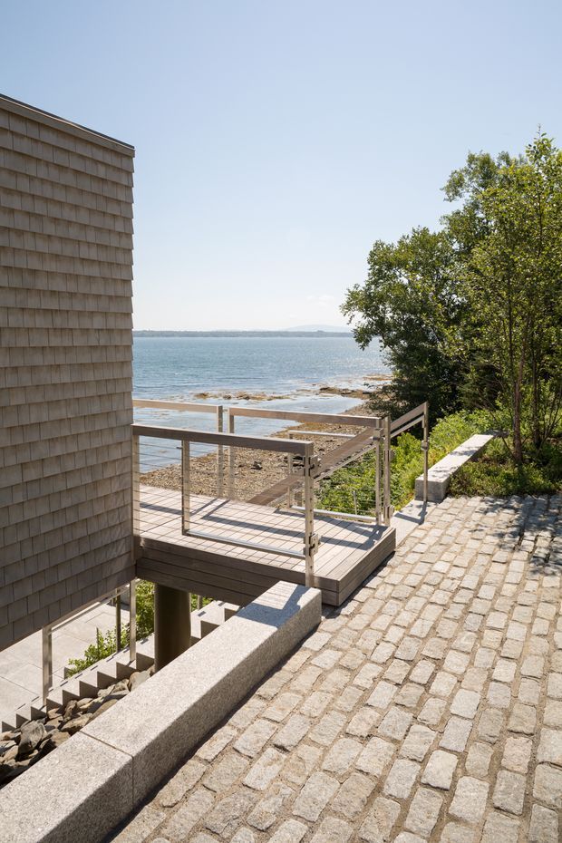 Exterior architectural view of a shingle-clad coastal building beside a rocky shoreline. A timber deck with minimalist metal-and-glass balustrades steps down toward the water, while a broad cobblestone path and concrete retaining walls define the landscaped approach. Bright clear daylight and expansive water views create a calm waterfront setting.