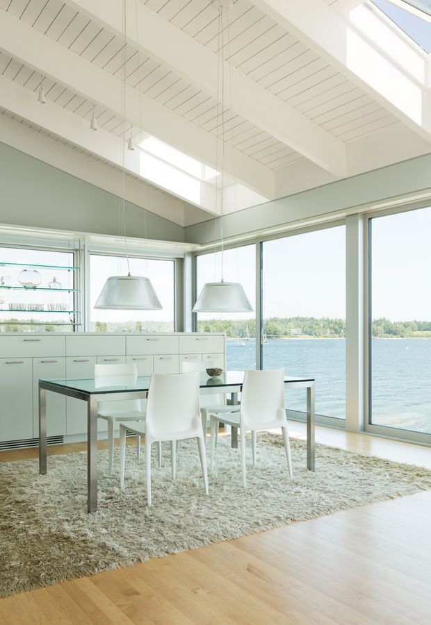 Bright, minimalist dining area beside expansive floor-to-ceiling glazing overlooking a body of water. The room features a white built-in storage and display wall, glass dining table with white chairs, pale timber flooring, a large textured neutral rug, suspended pendant lights, and a white slatted vaulted ceiling with skylights.