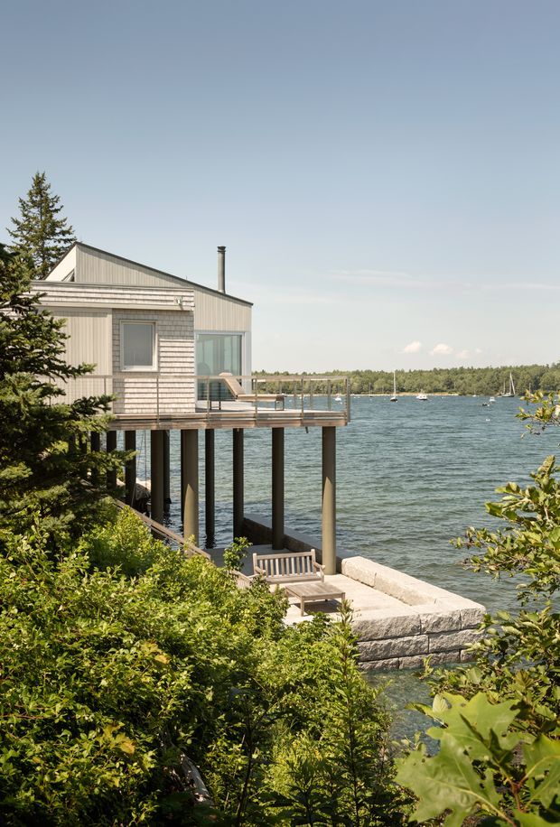 Exterior view of a light grey shingle-clad waterfront house projecting over a lake on dark structural stilts. The building has a steep gable roof, large glazed openings, a timber deck with railing, and a stone waterfront terrace and steps surrounded by dense greenery. Sailboats and a wooded shoreline are visible across the water.