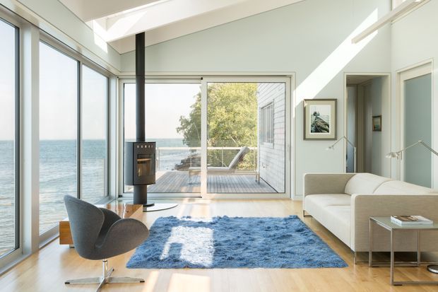 A bright contemporary living room overlooking the sea, with floor-to-ceiling glazing, a central freestanding wood-burning stove, pale timber flooring, a blue area rug, sculptural lounge chair, and cream sectional sofa. Sliding glass doors open to a timber deck, while skylights and high-level glazing bring in abundant daylight.
