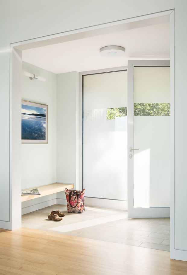 Bright, minimalist entryway with pale green walls, white trim, and a built-in bench with drawers. A frosted-glass exterior door and adjacent full-height frosted glazing provide privacy while admitting natural light. The floor transitions from light timber in the adjoining space to pale tile in the entry, with a framed landscape print and everyday items near the bench.