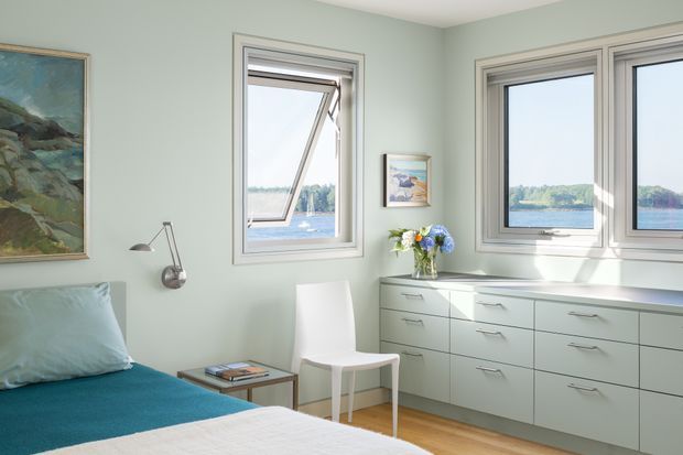 Bright, calm bedroom with pale green walls, a teal upholstered bed, minimalist white chair, and extensive built-in light grey cabinetry beneath multiple windows. The windows offer a view toward a body of water and wooded shoreline. A framed artwork, articulated wall lamp, small side table, and vase of flowers add restrained decorative touches.