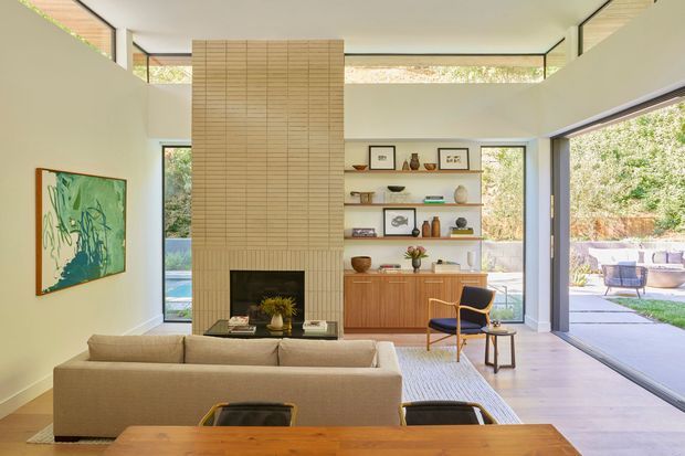 A bright mid-century-inspired living room and dining area with a tall tiled fireplace chimney as the focal point, built-in open shelving, and a timber sideboard. Large glazed openings connect the room to a landscaped outdoor patio. A neutral sofa, timber dining table, sculptural lounge chair, artwork, and warm wood finishes create an inviting, refined interior.