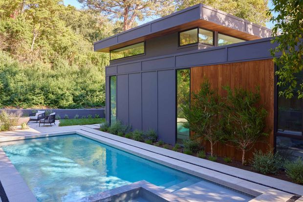 A contemporary flat-roof pool house or residential extension set within a wooded landscape. The compact building features dark grey exterior cladding, extensive horizontal clerestory-style glazing, warm timber soffits, and a prominent vertical timber feature wall. A long turquoise swimming pool with pale paving occupies the foreground, with a small outdoor seating area beside planted borders.