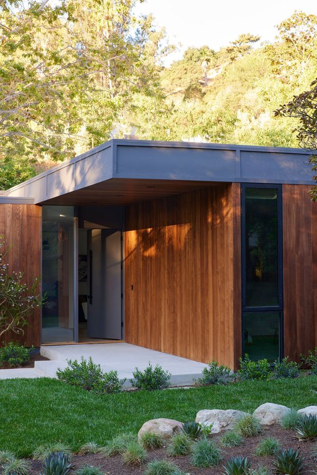 Exterior view of a compact contemporary flat-roofed house set within a lush landscaped garden. The facade combines warm vertical timber cladding with dark blue-grey painted fascia and slim dark-framed glazing. A recessed covered entry with a broad glass door opens onto a pale concrete path, while ornamental grasses, succulents, lawn and natural rocks surround the building.