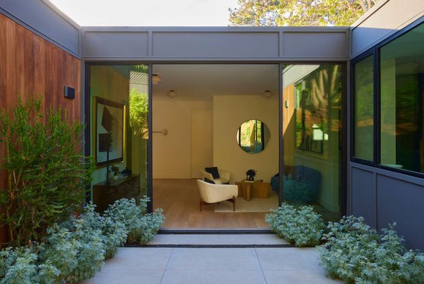 View from an outdoor courtyard toward a modern home's glazed entry and living area. The composition features dark painted metal window and door frames, warm timber wall cladding, extensive glazing, a paved path, and dense low planting. Inside, a small sitting area with a cream lounge chair, rug, timber side tables, artwork, and a round mirror is visible.