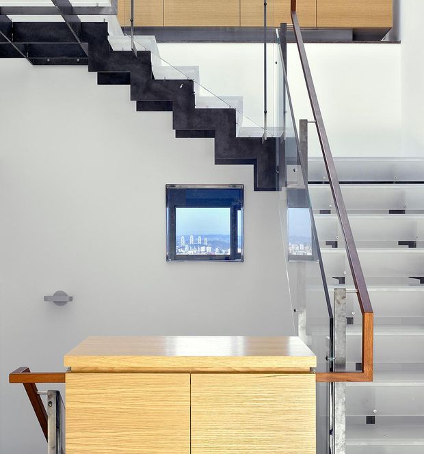 Interior view of a contemporary stairway and landing featuring a sculptural dark-finished floating staircase, clear glass balustrades, slim metal supports, and a timber handrail. Built-in light timber cabinetry occupies the lower foreground, while a small square window and bright white walls create a clean, modern composition.