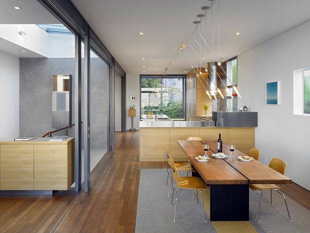 Open-plan contemporary dining and kitchen interior with a long timber dining table, mustard upholstered chairs, a dark central table base, light timber kitchen cabinetry, a grey stone island benchtop, timber flooring, and a striking cluster of suspended pendant lights. Full-height dark-framed glazing separates the dining area from an adjacent space and brings in abundant natural light.