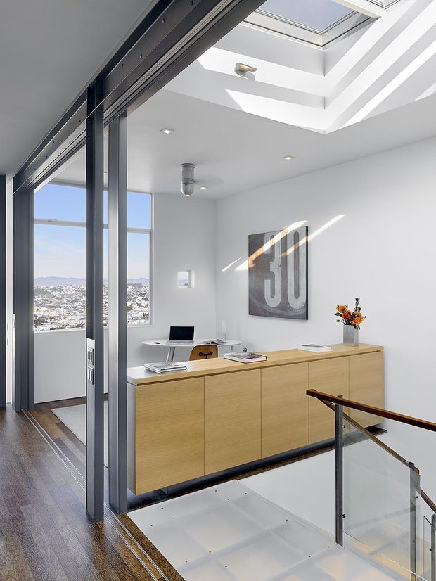 Bright contemporary home office on an upper-level landing, featuring a long light-wood built-in storage cabinet and desk, a small round work table, dark-framed internal glazing or sliding doors, polished timber flooring, and a glass-and-dark-metal stair balustrade. A large skylight and clerestory-like roof glazing flood the white room with daylight, while a cityscape is visible through the windows.