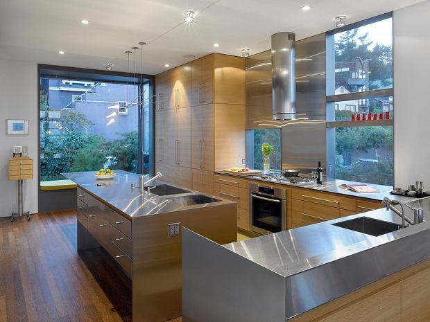 A spacious, high-end contemporary kitchen with extensive stainless-steel benchtops, two large islands, integrated appliances, warm timber cabinetry, and expansive corner glazing. A prominent cylindrical stainless-steel rangehood hangs above the cooktop, while pendant and recessed lighting illuminate the open, polished interior. The kitchen overlooks landscaped residential surroundings through large windows.